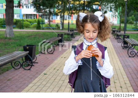 Girl with a backpack eating pie near school. A quick snack with a bun, unhealthy food, lunch from school. Back to school. Education, primary school classes, September 1 Girl with a backpack eating pie near school. A quick snack with a bun, unhealthy food, lunch from school. Back to school. Education, primary school classes, September 1 106312844