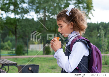 Girl with a backpack eating pie near school. A quick snack with a bun, unhealthy food, lunch from school. Back to school. Education, primary school classes, September 1 Girl with a backpack eating pie near school. A quick snack with a bun, unhealthy food, lunch from school. Back to school. Education, primary school classes, September 1 106312854