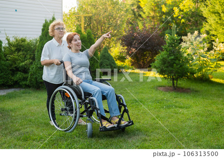 An elderly woman is carrying an adult daughter sitting in a wheelchair. Caucasian woman pointing her finger admiringly while walking outdoors. An elderly woman is carrying an adult daughter sitting in a wheelchair. Caucasian woman pointing her finger admiringly while walking outdoors. 106313500