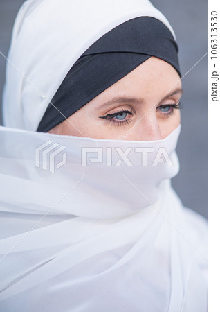 Portrait of a young blue-eyed woman in a hijab against a gray brick wall. Muslim woman with white scarf covering her face. Portrait of a young blue-eyed woman in a hijab against a gray brick wall. Muslim woman with white scarf covering her face. 106313530
