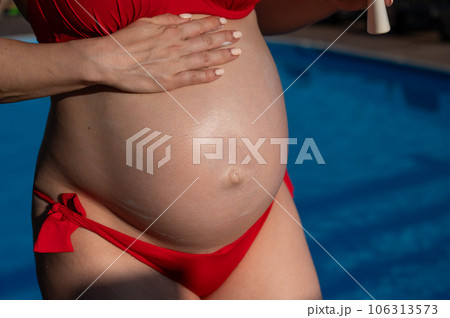 Close-up of the belly of a pregnant woman sunbathing in a red bikini. Expectant mother applies sunscreen. Close-up of the belly of a pregnant woman sunbathing in a red bikini. Expectant mother applies sunscreen. 106313573