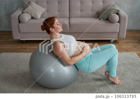 Pregnant red-haired woman doing arm exercises with fitball at home. Pregnant red-haired woman doing arm exercises with fitball at home. 106313785