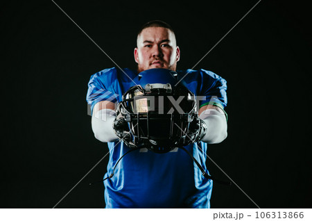 An Asian man with a red beard in a blue american football uniform holds a helmet on a black background. 106313866