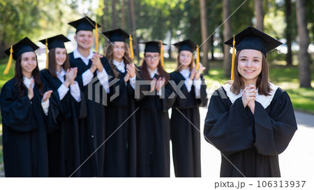 Group of happy students in graduation gowns outdoors. A young girl in the foreground. Group of happy students in graduation gowns outdoors. A young girl in the foreground. 106313937