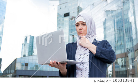 Thoughtful business woman in hijab and suit is holding a laptop outdoors. 106313941
