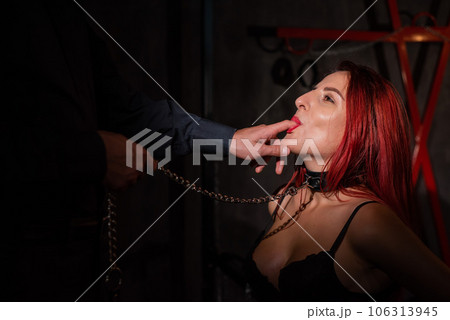 BDSM. Portrait of a red-haired woman in shackles kneeling and licking the fingers of a man. BDSM. Portrait of a red-haired woman in shackles kneeling and licking the fingers of a man. 106313945