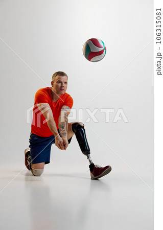 Confident man with prosthetic legs, disability in uniform hits volleyball ball in motion on floor. Inclusive sport for people with disabilities Confident man with prosthetic legs, disability in uniform hits volleyball ball in motion on floor. Inclusive sport for people with disabilities 106315081
