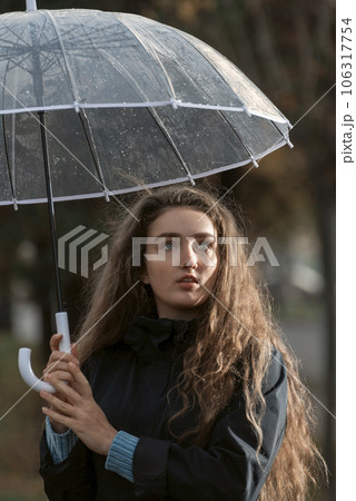 Blue-eyed girl with wavy hair looks wistfully to side holding transparent umbrella in hands. Vertical frame. Blue-eyed girl with wavy hair looks wistfully to side holding transparent umbrella in hands. Vertical frame. 106317754