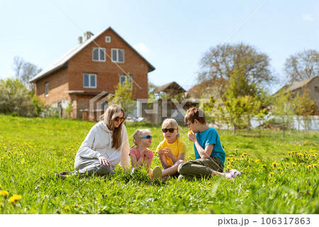 Mom or nanny sitting with children in meadow surrounded by dandelions. Young family sitting in the grass against two-story house. 106317863