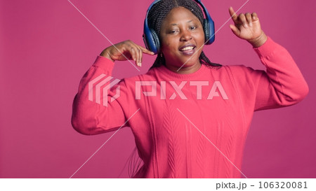 Joyful black woman wearing modern earphones moving around and having a good time. Lady with african american ethnicity confidently dancing to good music from stereo headphones. 106320081