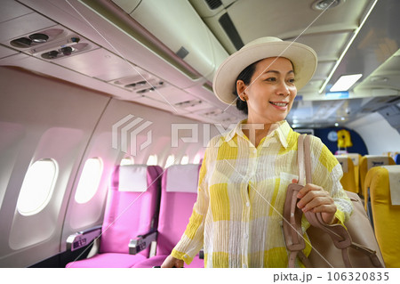 Smiling 60s female tourist with suitcase walking the aisle on plane. Travel and summer vacation concept 106320835