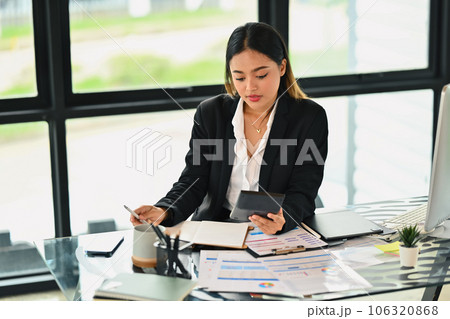 Focused millennial female financier analyzing marketing data and using calculator at office desk Focused millennial female financier analyzing marketing data and using calculator at office desk 106320868