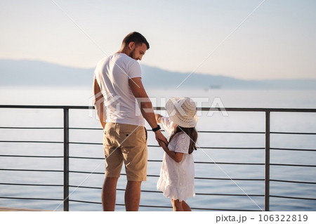 Back view of daughter with her father stand on heigh balcony in front of sea sunset. Dad with his little girl looking on mountains Back view of daughter with her father stand on heigh balcony in front of sea sunset. Dad with his little girl looking on mountains 106322819