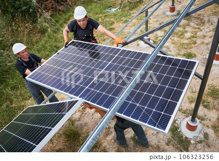 Workers mounting photovoltaic solar panel system outdoors. Men engineers placing solar module on metal rails, wearing construction helmets and work gloves. Renewable and ecological energy. Workers mounting photovoltaic solar panel system outdoors. Men engineers placing solar module on metal rails, wearing construction helmets and work gloves. Renewable and ecological energy. 106323256