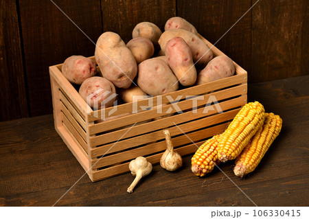 wooden box full of potatoes, garlic bulbs and corn cobs on dark wooden background 106330415