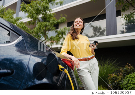 Close up of beautiful woman using smartrphone while charging her electric car on the street. 106330957