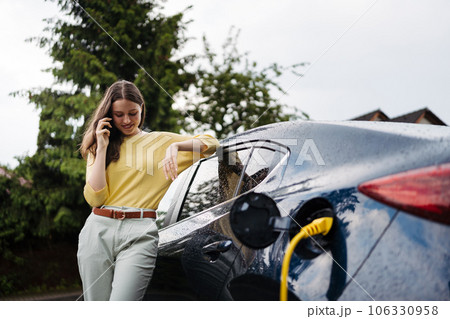 Close up of beautiful woman calling while charging her electric car on the street. Close up of beautiful woman calling while charging her electric car on the street. 106330958