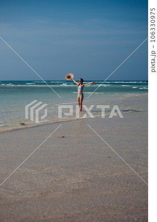 Portrait of slim woman standing on the beach in a bikini and a straw hat in hand. 106330975