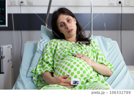 Pregnant patient wearing medical oximeter, looking at camera while waiting to receive medical examination from doctor. Woman expecting child and preparing to give birth in hospital maternity room Pregnant patient wearing medical oximeter, looking at camera while waiting to receive medical examination from doctor. Woman expecting child and preparing to give birth in hospital maternity room 106332789