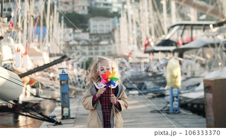 Little girl blows on a toy rainbow windmill by the sailboats in marina. Clean wind energy and peace concepts Little girl blows on a toy rainbow windmill by the sailboats in marina. Clean wind energy and peace concepts 106333370