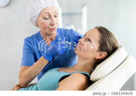 Mature woman doctor gives injection and performs procedure of lip augmentation of female patient 106334910