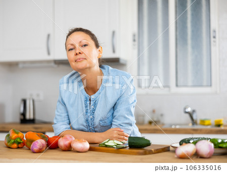 Elegant female in a blue blouse standing behind tabletop with fresh vegetables at the kitchen Elegant female in a blue blouse standing behind tabletop with fresh vegetables at the kitchen 106335016