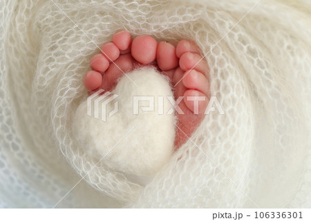 Knitted white heart in the legs of a baby. The tiny foot of a newborn baby. Soft feet of a new born in a white wool blanket. Close up of toes, heels and feet of a newborn. Macro photography 106336301