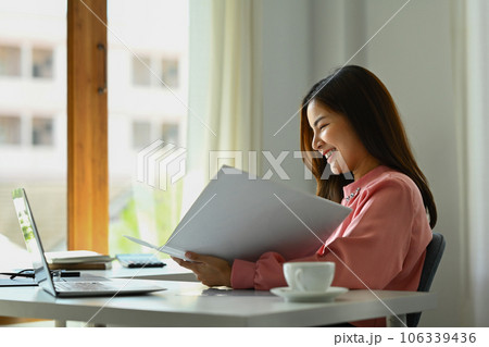 Smiling asian woman economist reading paper document, analyzing marketing data, statistics, financial results at desk Smiling asian woman economist reading paper document, analyzing marketing data, statistics, financial results at desk 106339436
