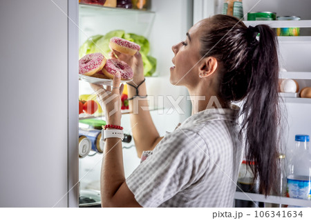 A young hungry woman is about to eat sweet donuts by the open refrigerator late in the evening 106341634