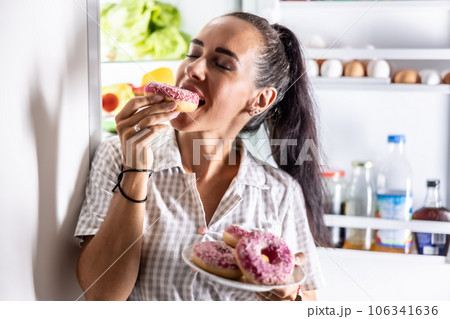 Hungry brunette in pajamas enjoys sweet donuts late at night by the open refrigerator 106341636