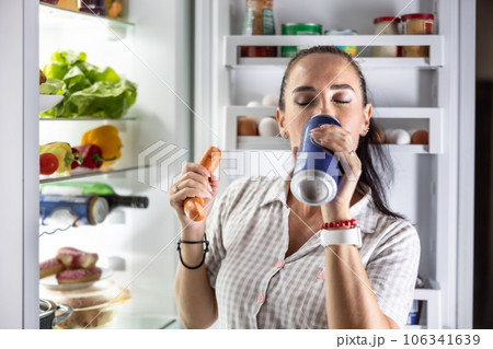 Hungry woman in pajamas drinks beer and eats sausages by the refrigerator at night Hungry woman in pajamas drinks beer and eats sausages by the refrigerator at night 106341639