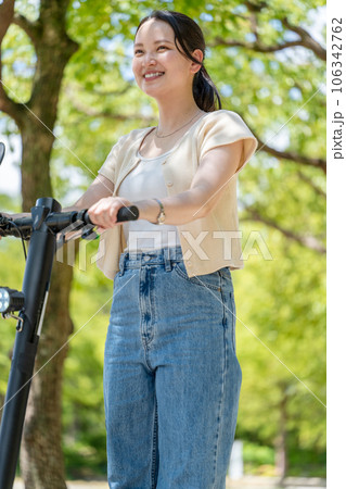 爽やかな陽射しと緑の中を電動キックボードで爽やかに走る若くてかわいい女性｜電動キックボードイメージ 106342762