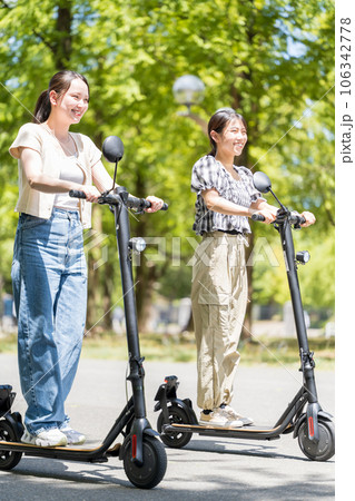爽やかな陽射しと緑の中を電動キックボードで爽やかに走る若くてかわいい女性｜電動キックボードイメージ 106342778