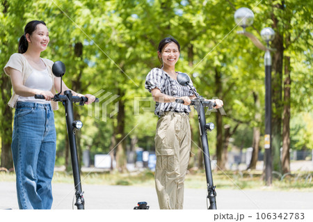 爽やかな陽射しと緑の中を電動キックボードで爽やかに走る若くてかわいい女性｜電動キックボードイメージ 106342783
