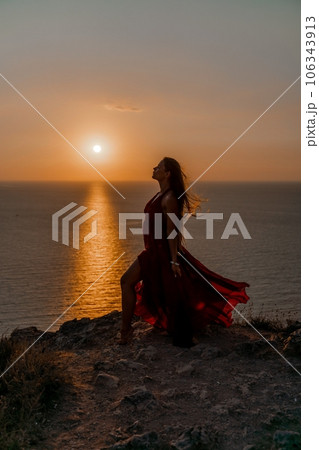 Woman sunset sea red dress, back view a happy beautiful sensual woman in a red long dress posing on a rock high above the sea on sunset. 106343913
