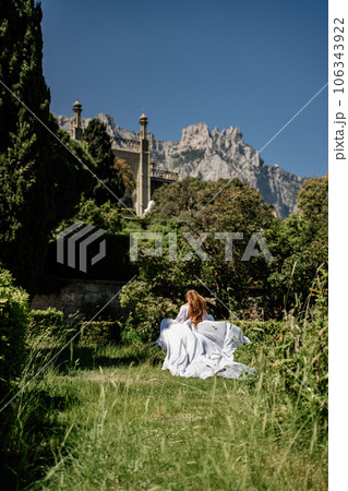 a beautiful woman with long brown hair and long white dress stay along a path along beautiful bushes in the park rear view 106343922