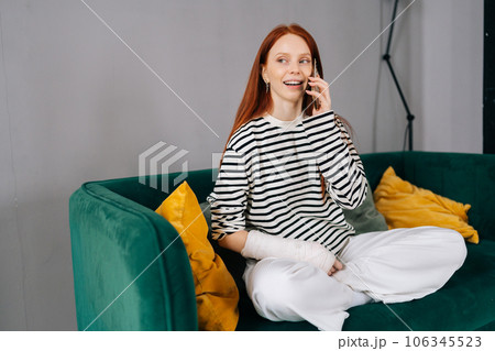 Front view of happy young woman with broken arm wrapped in gypsum bandage talking on smartphone with friends during rehabilitation, looking away. Smiling female with injured hand from accident at home 106345523