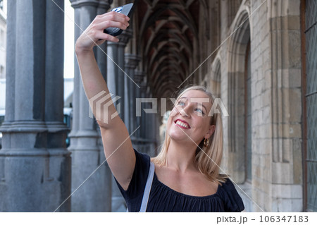 Beautiful young blonde Caucasian millenial woman taking a selfie with smartphone outdoors in a 106347183