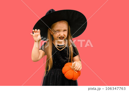 Portrait of winsome little witch smiling girl wearing a wizard hat and dressed in stylish carnival dress, looking at camera posing against pink background, Halloween 106347763