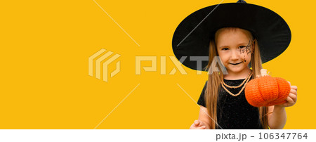 Banner with portrait of a little girl in a witch costume with a spider web pattern on his face and a pumpkin in his hands on the orange background. Copy space. 106347764