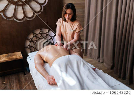 High-angle view of unrecognizable man getting back massage in spa salon lying on massage table from professional masseuse. Male client lying on stomach having full body treatment in wellness center. 106348226
