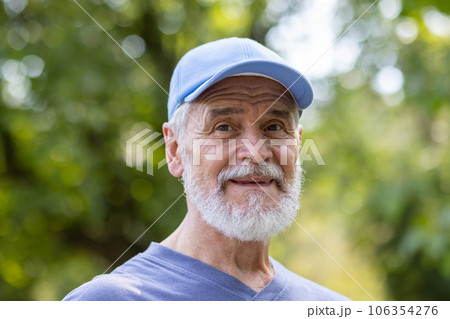 Portrait of senior mature gray-haired man in park, pensioner in sports cap smiling and looking at camera close-up among trees, active physical exercise and active lifestyle. 106354276