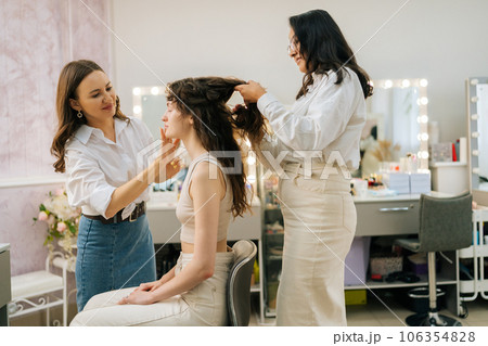 Side view of positive female make up artist and hairdresser prepare to doing makeup and hairstyle for young woman in beauty salon sitting in front of mirror. Four-handed makeup and styling concept. Side view of positive female make up artist and hairdresser prepare to doing makeup and hairstyle for young woman in beauty salon sitting in front of mirror. Four-handed makeup and styling concept. 106354828