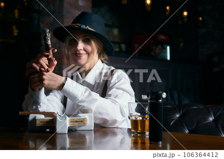Senior attractive female gangster smoking cigar over dark background in restaurant Senior attractive female gangster smoking cigar over dark background in restaurant 106364412