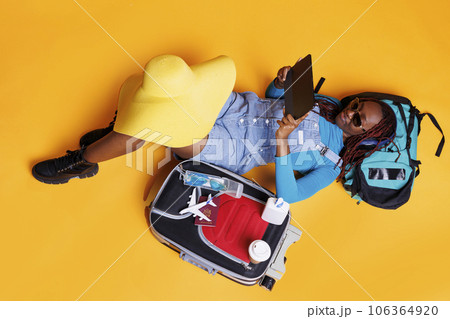 Young person watching film on tablet, sitting on the floor and waiting for flight departure on camera. Female traveller looking at movie on device, preparing to leave on vacation trip. Young person watching film on tablet, sitting on the floor and waiting for flight departure on camera. Female traveller looking at movie on device, preparing to leave on vacation trip. 106364920
