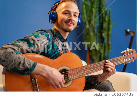Young man with headphones sitting on sofa and playing guitar 106367438
