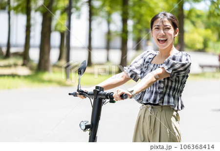 輝く陽射しと緑の木々の中を電動キックボードで爽やかに走る若くてかわいい女性｜電動キックボードイメージ 106368414