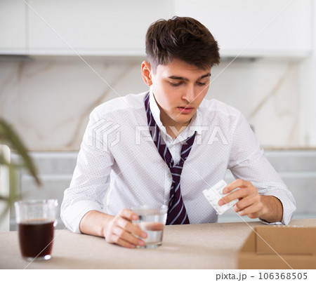 Sick young man with hangover drinks pills while sitting at table 106368505