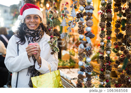 Cheerful hispanic woman in Santa hat with cup of hot tea walking at Christmas street fair 106368508
