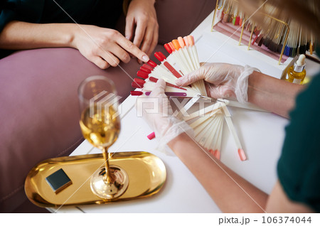Close up of female hand pointing at red color nail sample while choosing manicure with manicurist. Manicure master in sterile gloves holding nail polish color palette. 106374044
